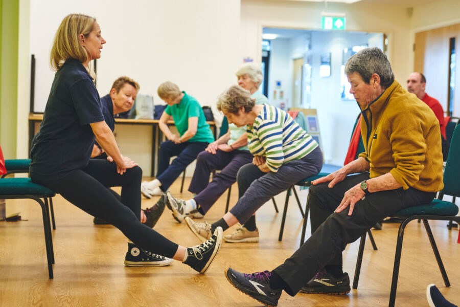 Older participants doing seated ham string stretches led by an iCareiMove female instructor.