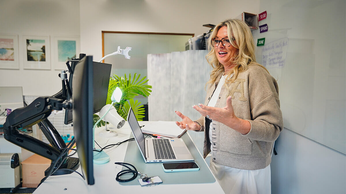 iCareiMove Helen Tite speaking to their computer on a video call in a light office with houseplants.