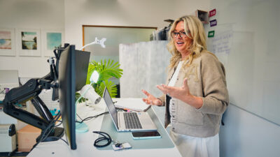 iCareiMove Helen Tite speaking to their computer on a video call in a light office with houseplants.
